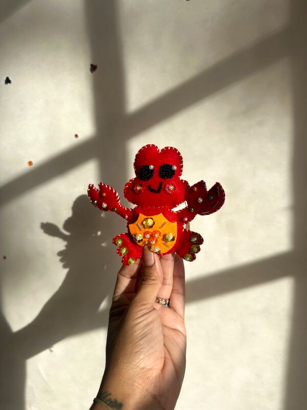 Mano sosteniendo un cangrejito rojo con barriga naranja, sonrisa bordada y piedritas.