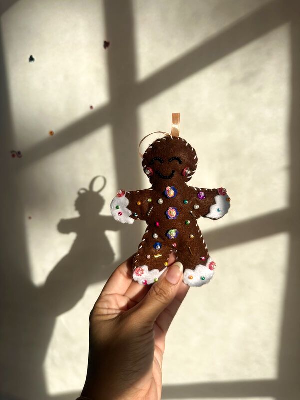 Mano sosteniendo una galleta de jengibre café con sonrisa bordada, botones de lentejuela y bordes blancos como glaseado.