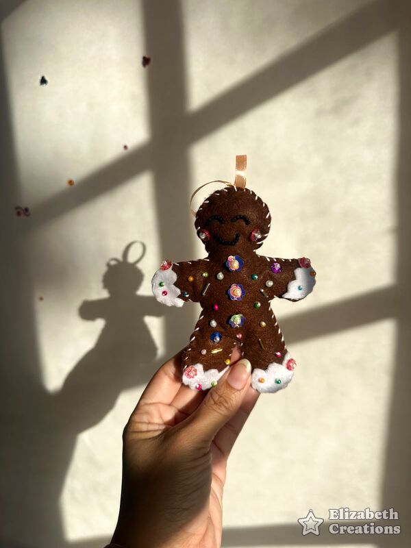 Mano sosteniendo una galleta de jengibre café con sonrisa bordada, botones de lentejuela y bordes blancos como glaseado.