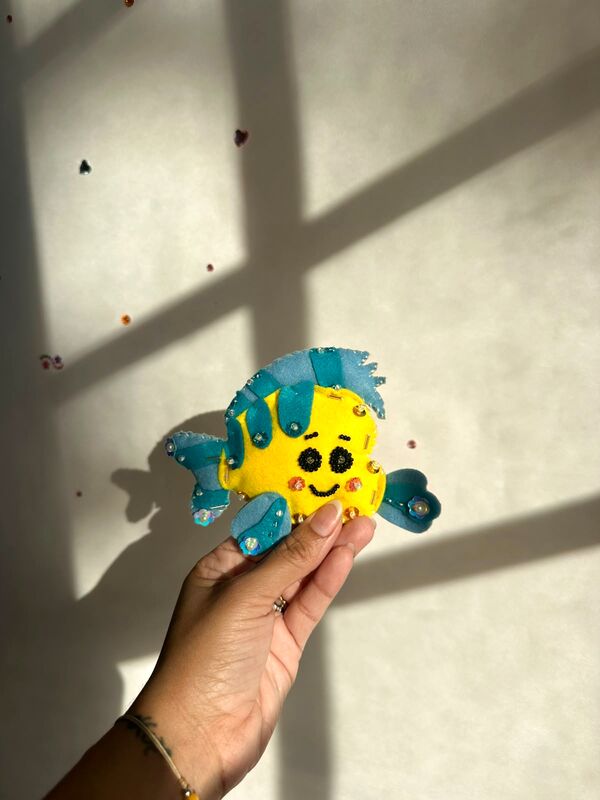 Mano sosteniendo un pececito amarillo con aletas turquesa y ojos bordados con perlitas negras.