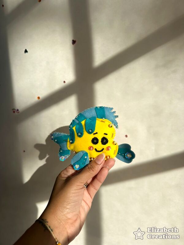 Mano sosteniendo un pececito amarillo con aletas turquesa y ojos bordados con perlitas negras.