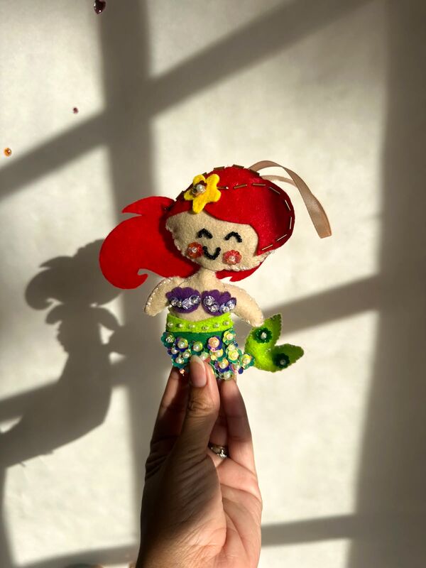 Mano sosteniendo una sirenita de pelo rojo, con cola verde llena de lentejuelas.