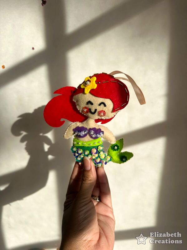 Mano sosteniendo una sirenita de pelo rojo, con cola verde llena de lentejuelas.