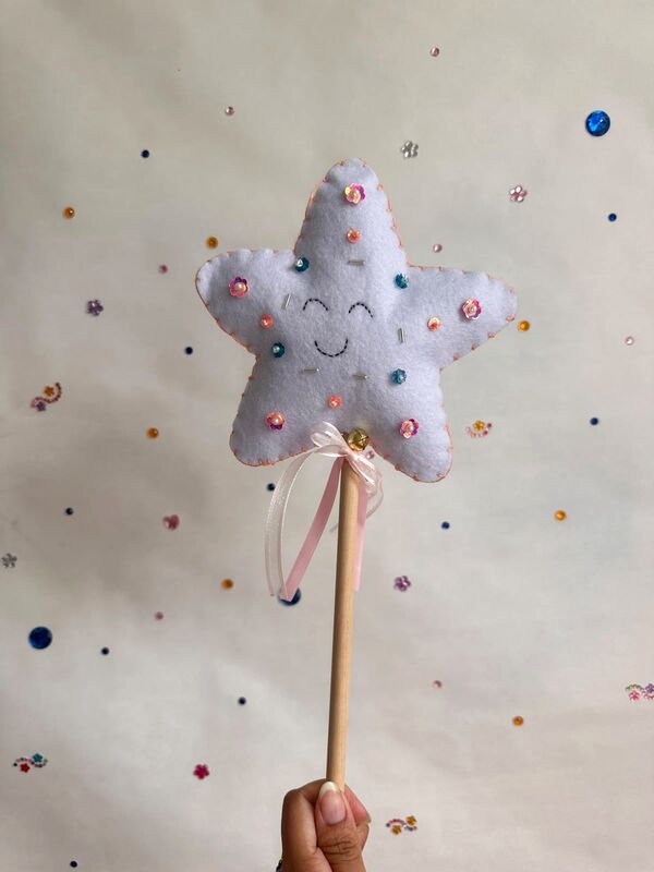 Mano sosteniendo una varita mágica: una estrella blanca con carita sonriente bordada, lentejuelas de colores, cinta rosada y un cascabel, en un palito de madera.