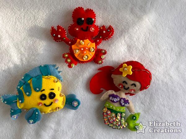 El set Bajo el mar sobre una cobija blanca: un cangrejito rojo, un pececito amarillo con aletas turquesa y una sirenita de pelo rojo, con sus caritas bordadas.