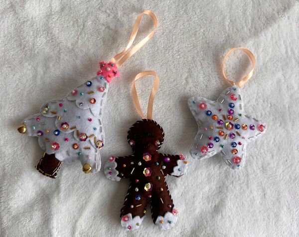 El set Navidad nevada sobre una cobija blanca: un arbolito blanco con estrella rosada, una galleta de jengibre y una estrella blanca, con lentejuelas de colores y cintas color durazno para colgar.
