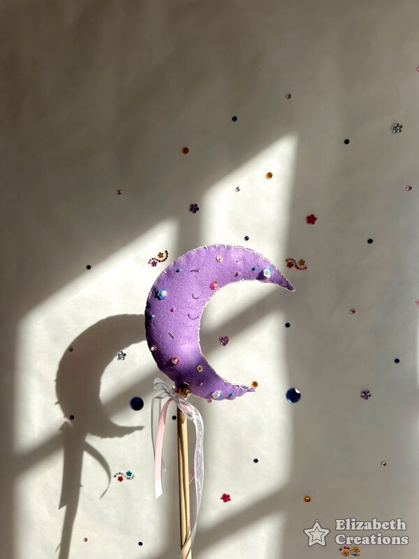 Varita mágica: una luna lila con perlitas, en un palito de madera con cintas, a la luz de la ventana.